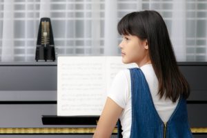 Young girl in a denim dress looking away from a piano with adult piano lessons sheet music on the stand.